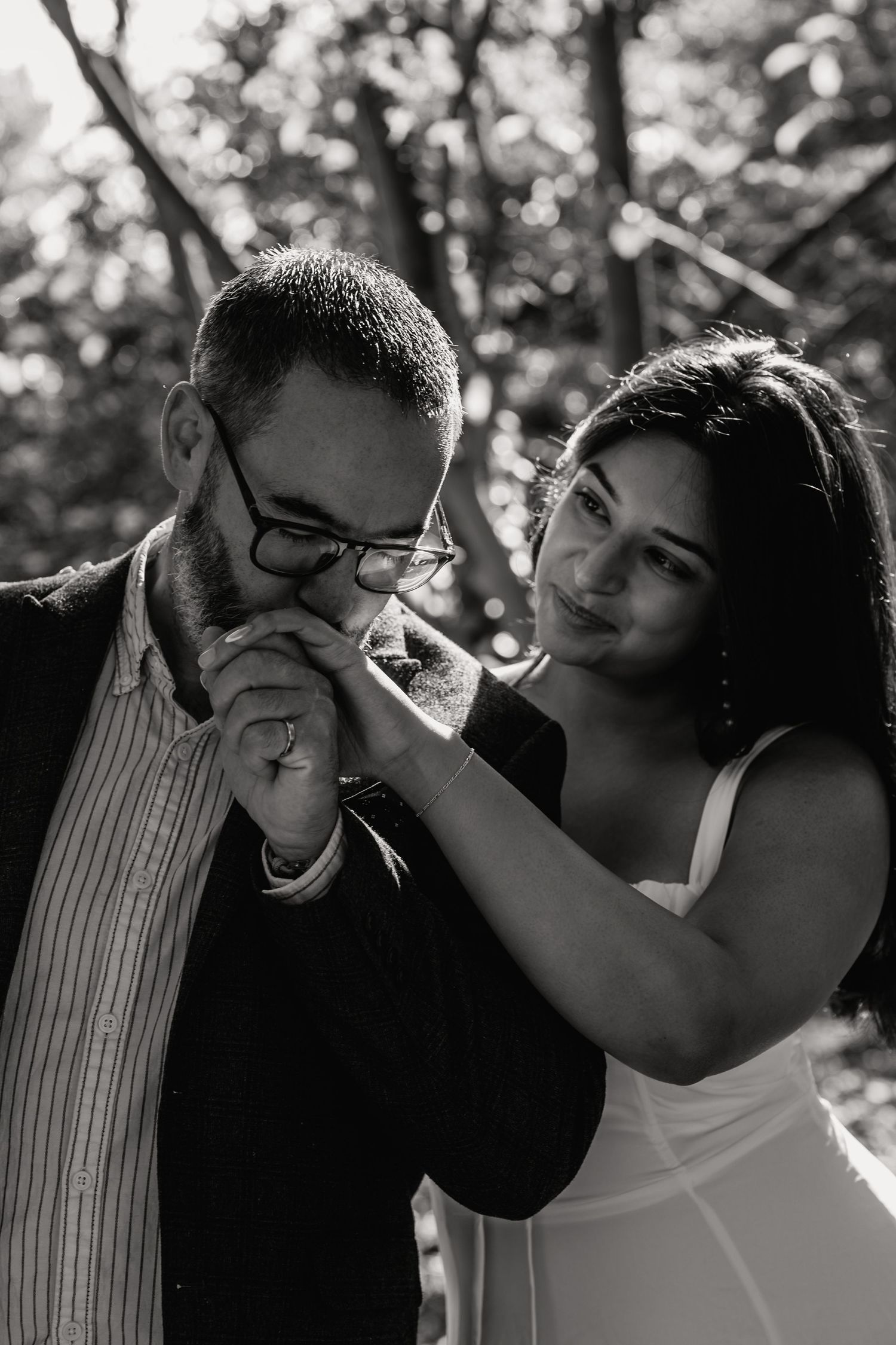 Black and white photo of a man in glasses kissing a woman's hand outdoors, romantic couple portrait.