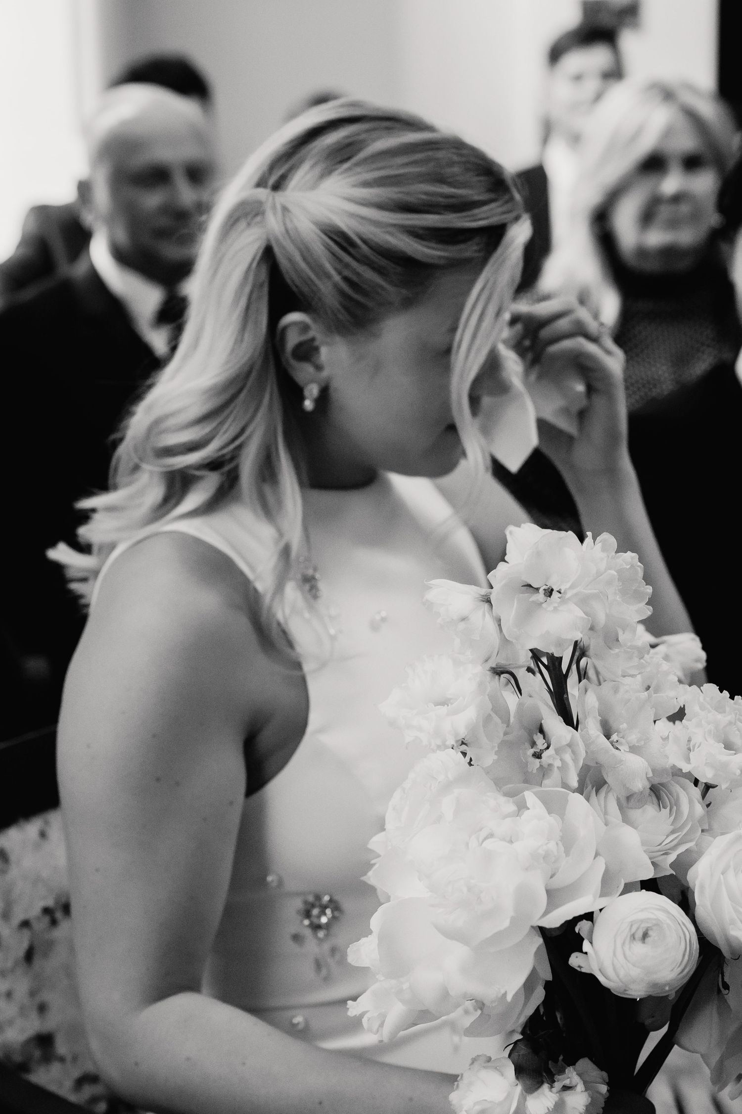 Emotional bride in white gown wiping tears holding large white floral bouquet in black and white.
