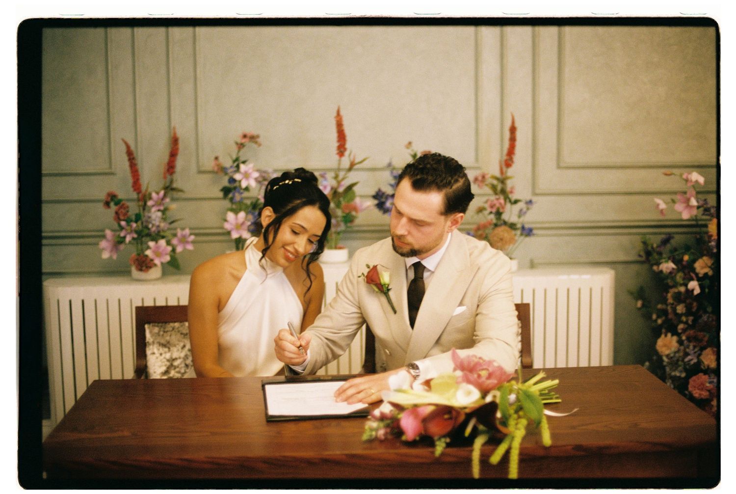 Bride and groom sign the marriage register together at a decorated table with floral arrangements.