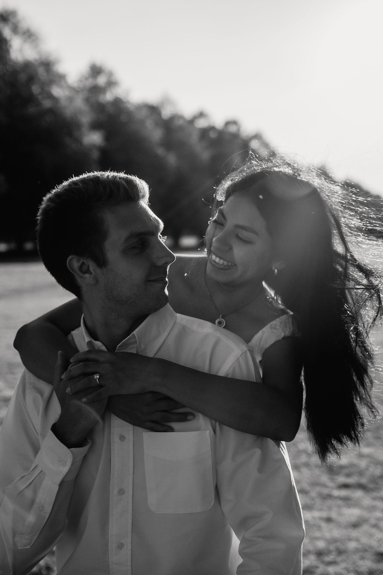 Black and white photo of smiling couple embracing outdoors with sunlight streaming through trees behind them.