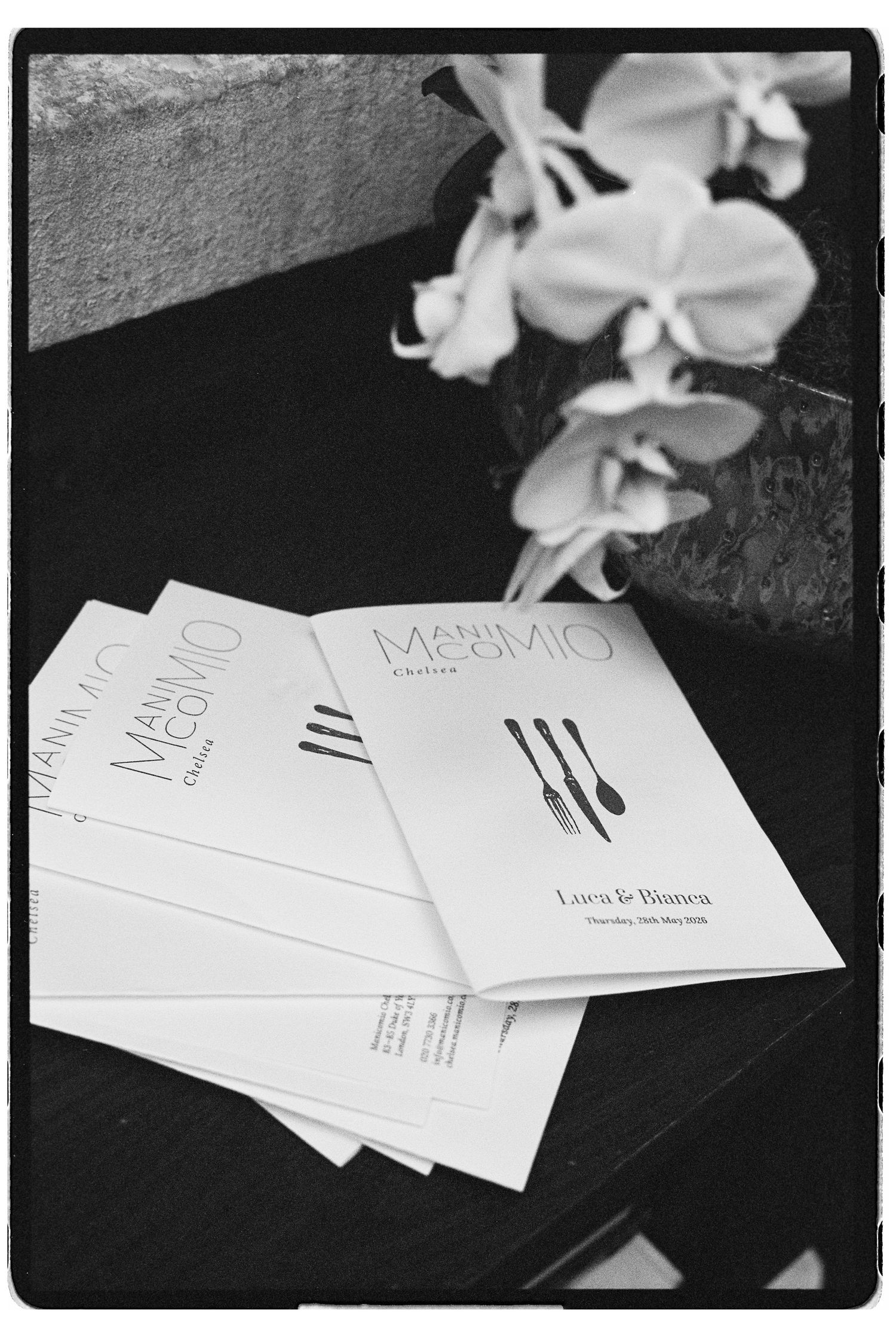 Black and white photo of Manu Mio Chelsea menus for Luca and Bianca beside white orchids.