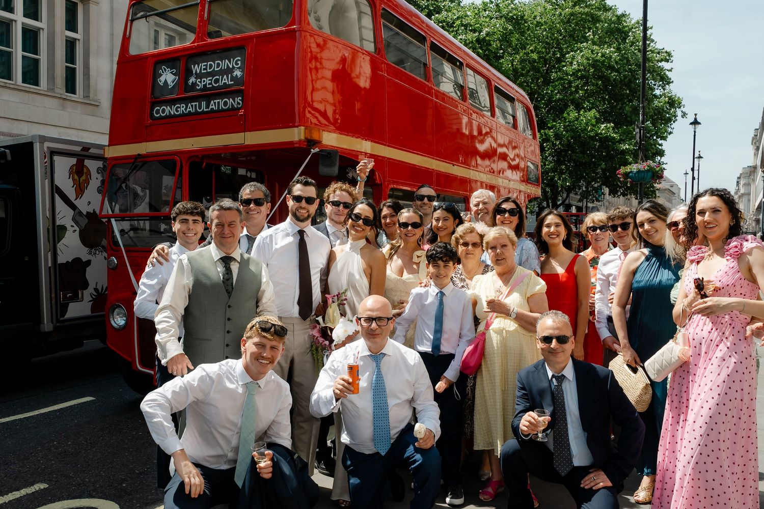 Wedding party group photo in front of a red double-decker London wedding bus on a sunny day.