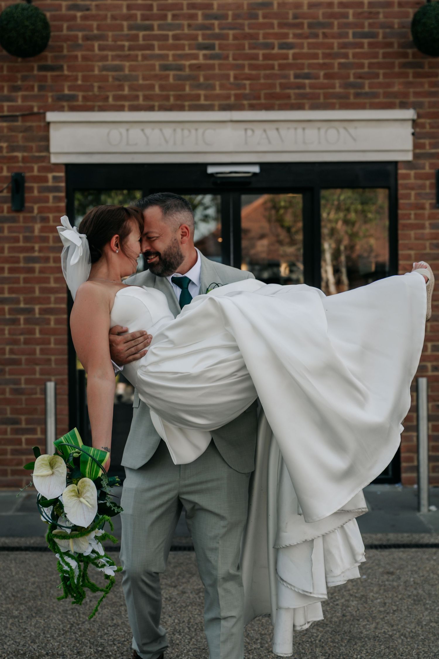 Groom carries bride in white gown outside Olympic Pavilion on their wedding day.