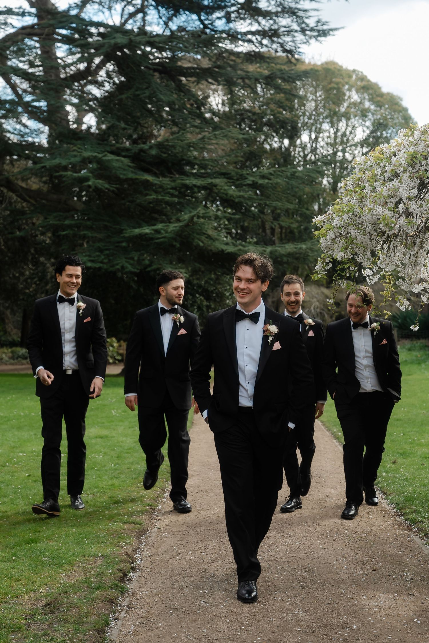 Groom and groomsmen in black tuxedos walking along a garden path at a wedding venue.