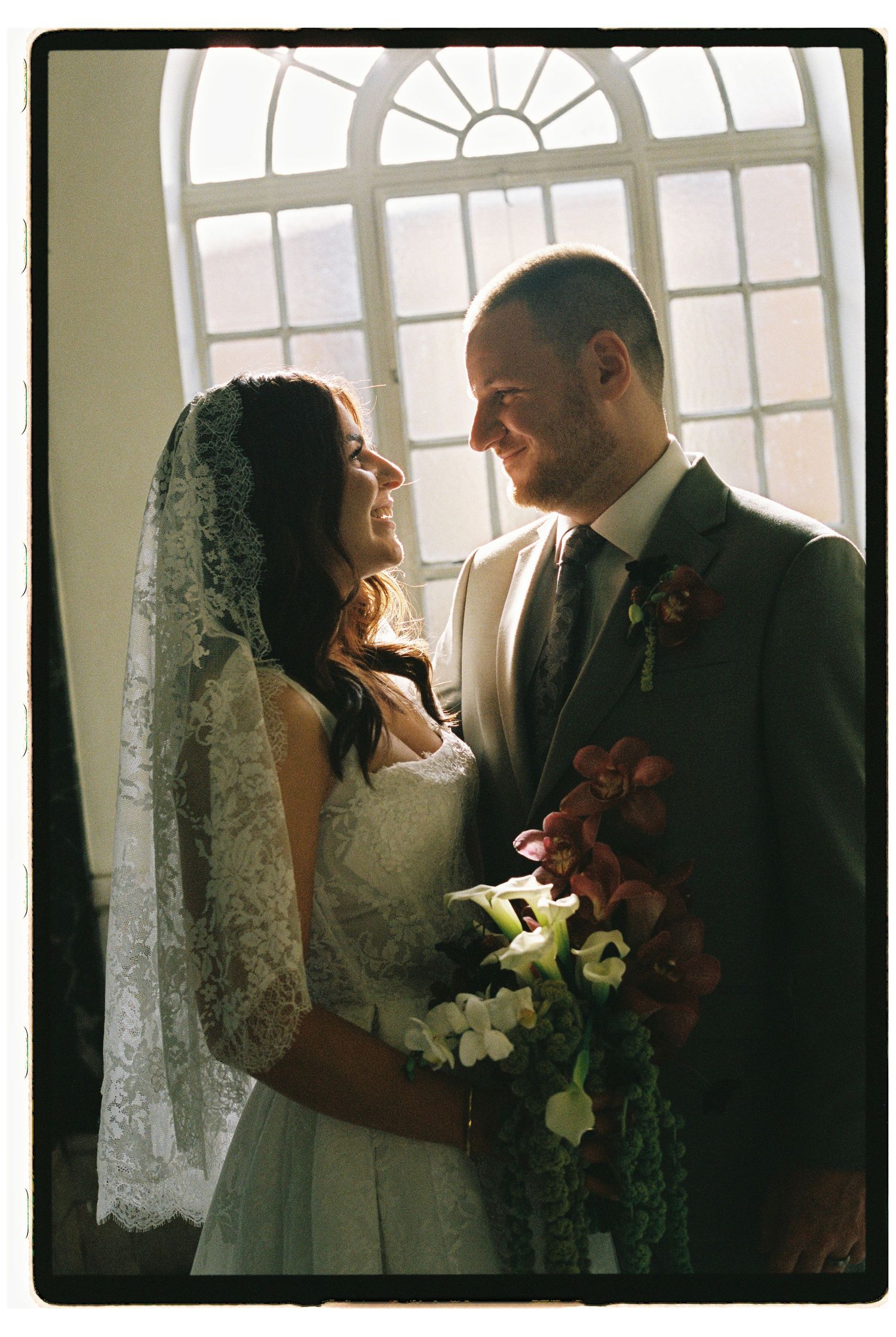 Bride and groom share a romantic moment by a large window, bride holding burgundy floral bouquet wearing lace veil.
