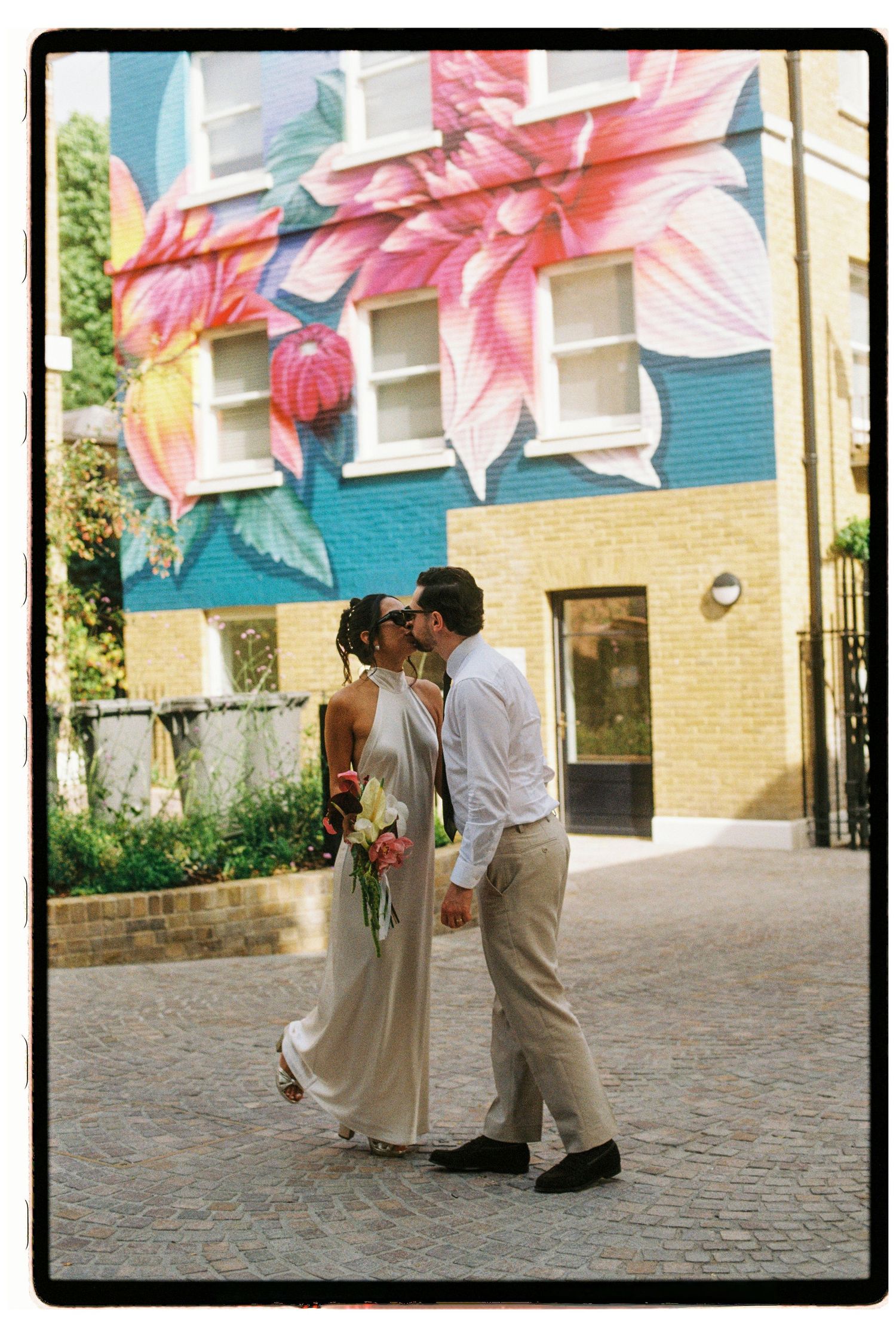 Bride and groom kissing in front of vibrant large floral mural on colorful building exterior.