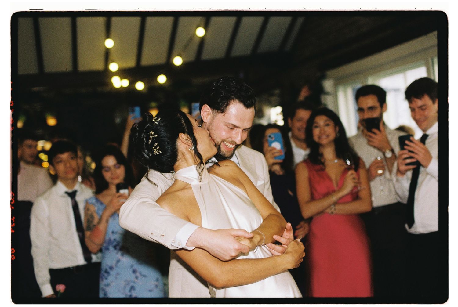 Bride and groom share first dance while wedding guests celebrate and photograph the moment in warmly lit reception venue.