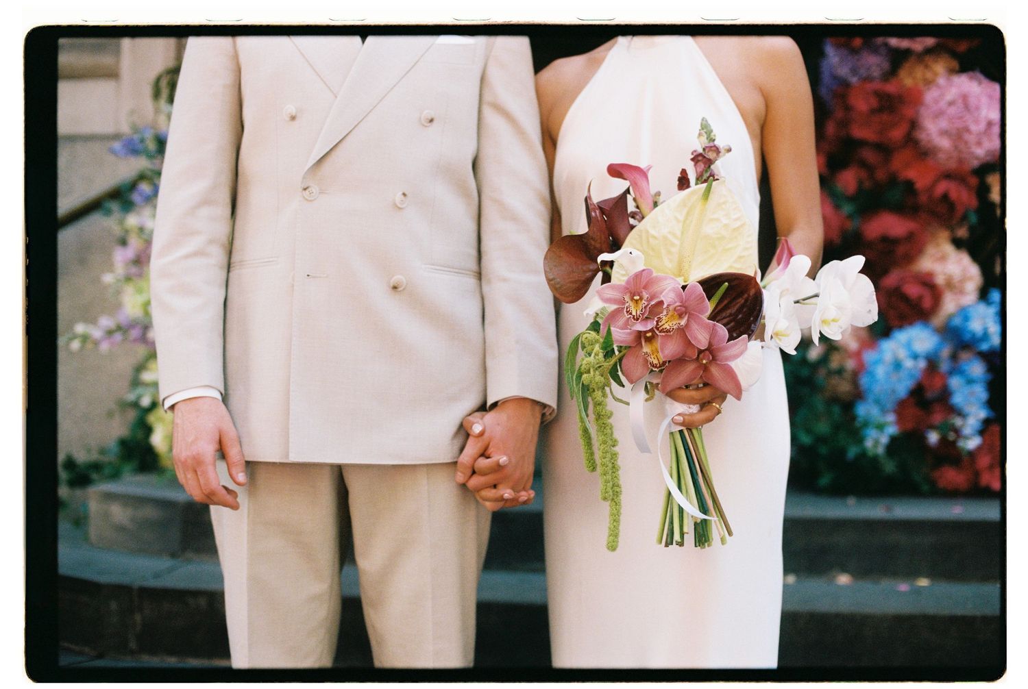 Bride and groom holding hands, bride holding colorful bouquet in white halter dress, groom in beige double-breasted suit.
