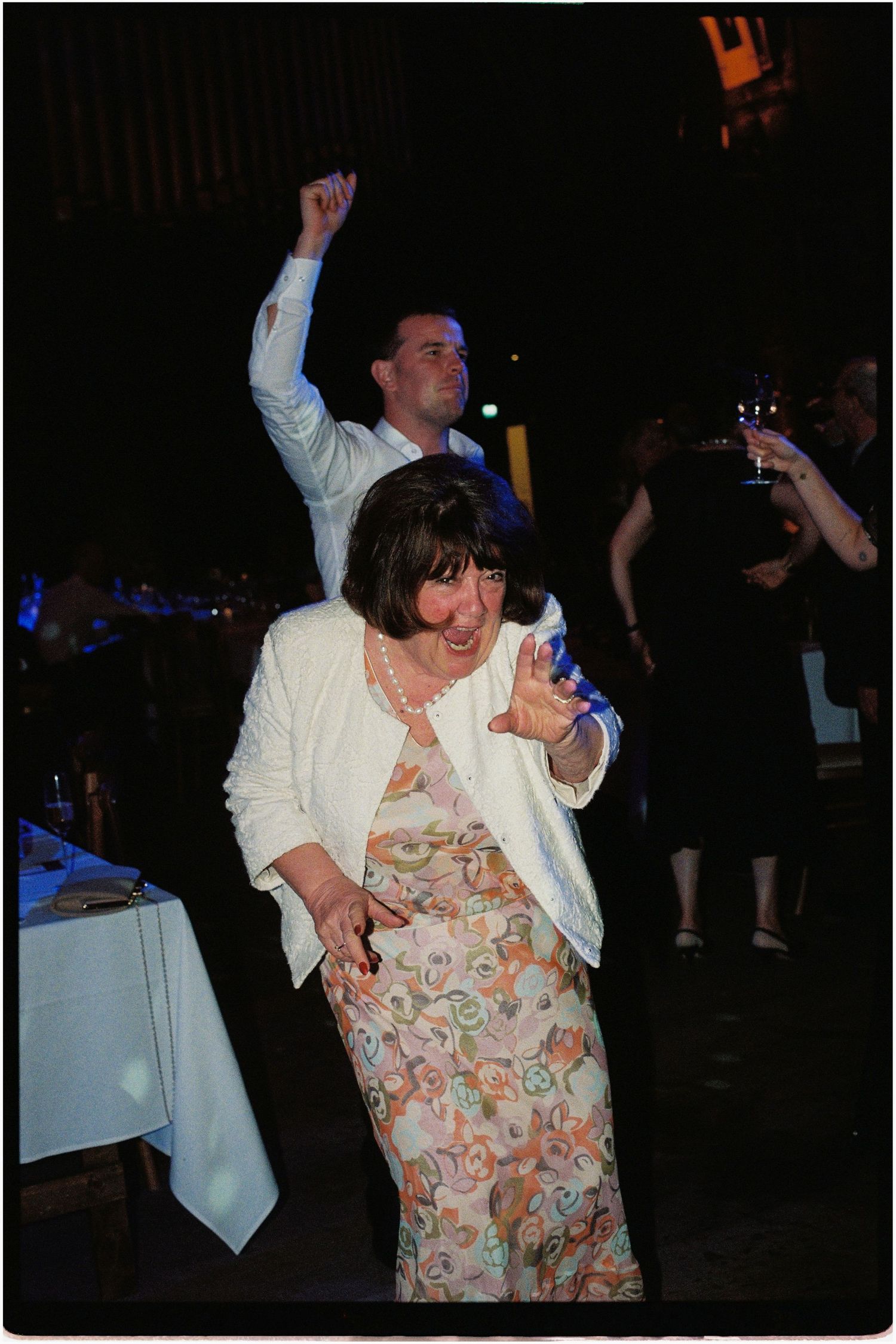 Elderly woman in floral dress and white blazer dancing joyfully at a wedding reception.