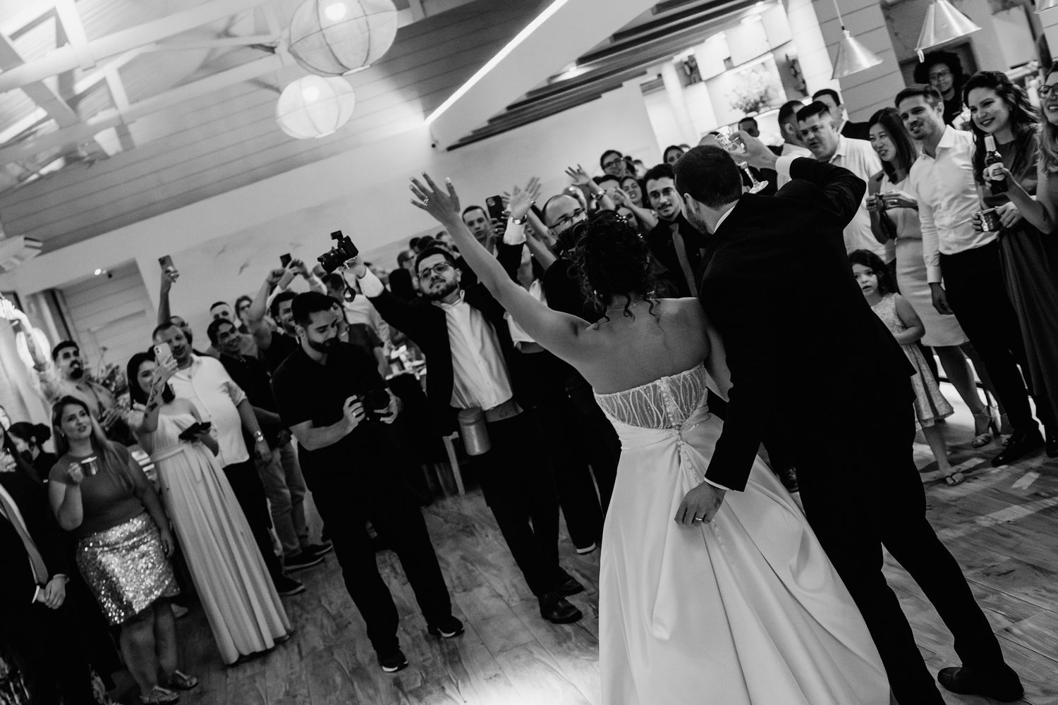 Bride and groom share first dance surrounded by cheering wedding guests in black and white photo.