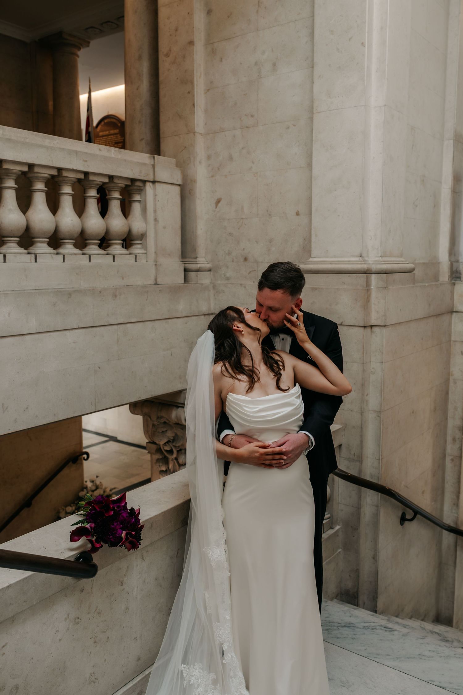 Bride and groom share a romantic kiss on marble staircase in elegant wedding venue with stone columns.