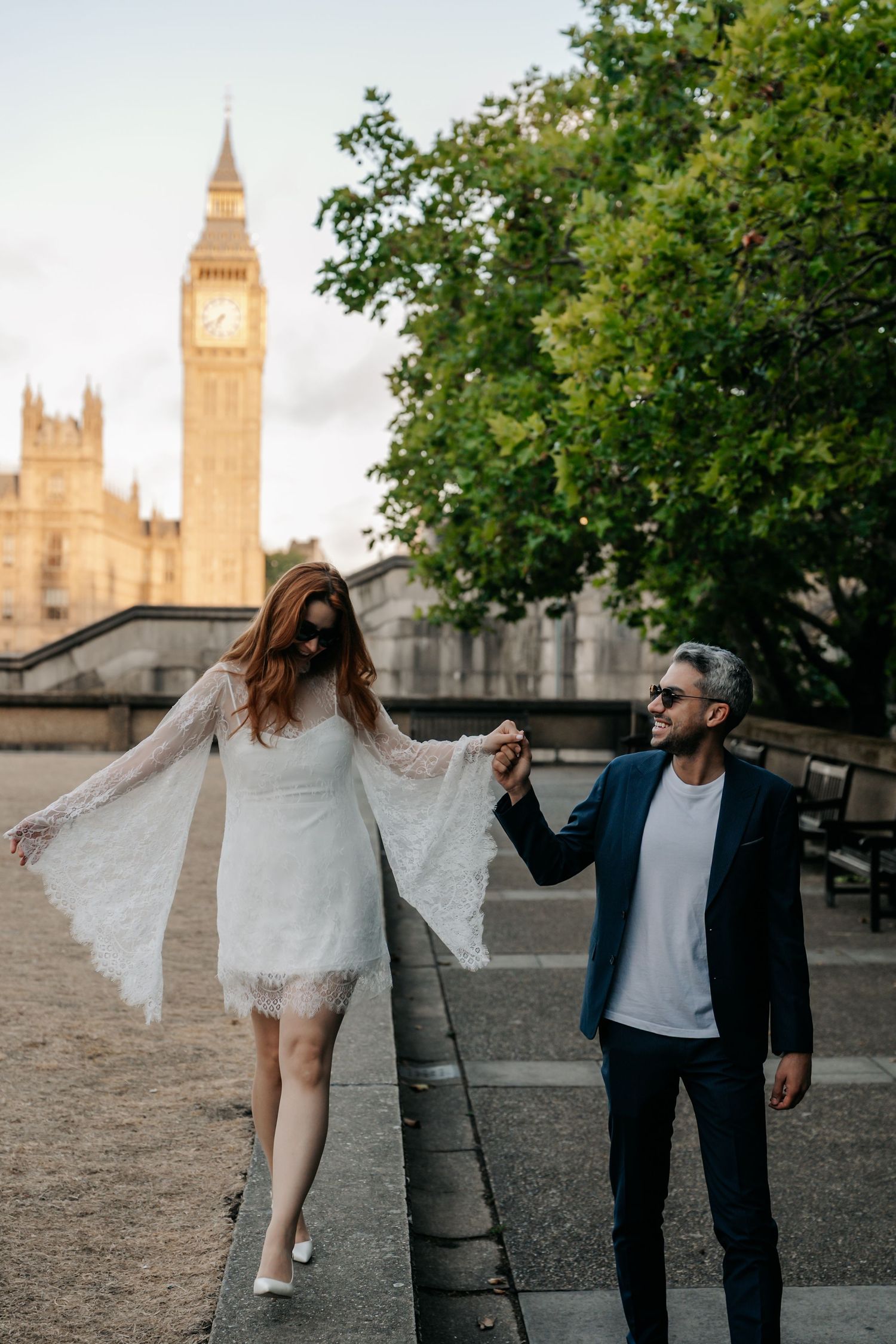 Couple holding hands in wedding attire walking near Big Ben, London, with the woman in a white lace dress and man in a navy suit.