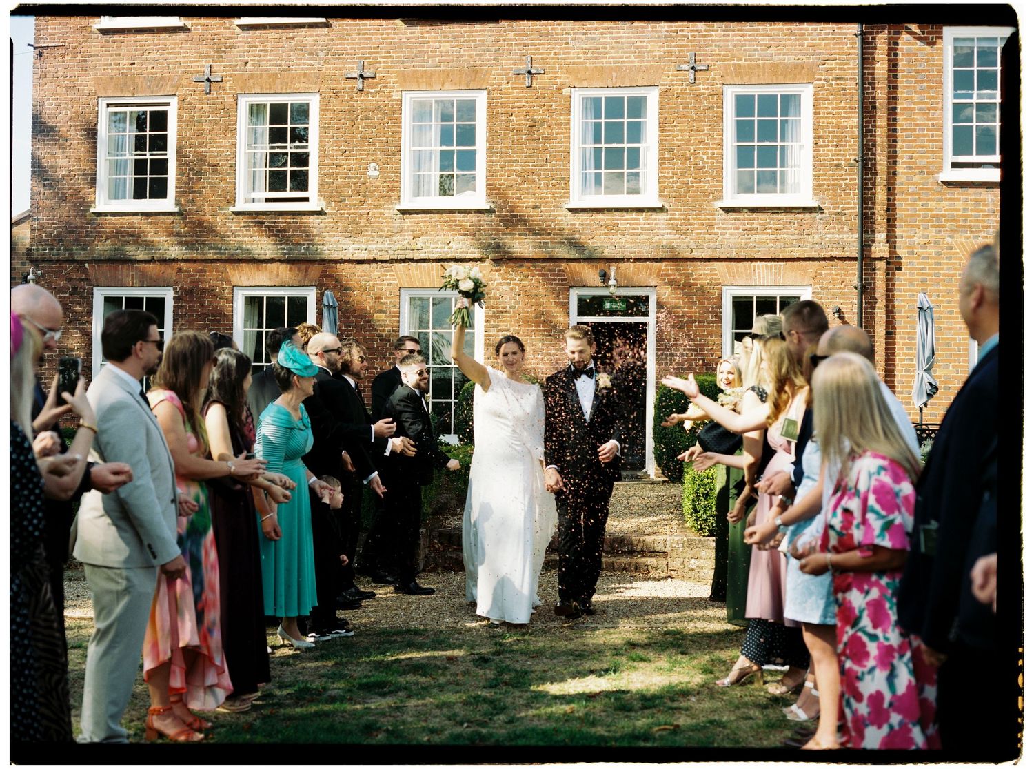 Bride and groom walk through confetti shower outside brick building, surrounded by cheering wedding guests.