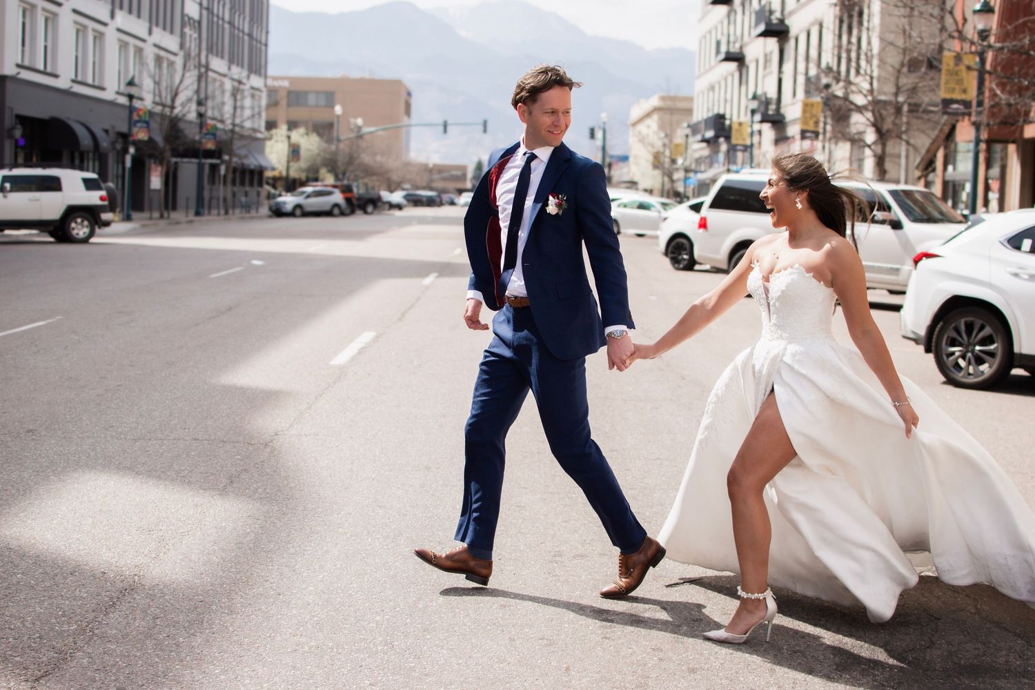 Bride and groom crossing a city street, bride in flowing white dress holding groom's hand in navy suit.