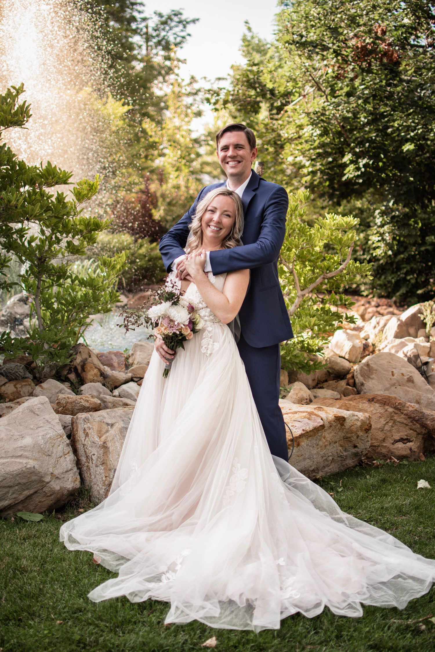 Bride and groom embrace in a lush garden setting, she holds a floral bouquet in a white gown as he wears a navy suit.