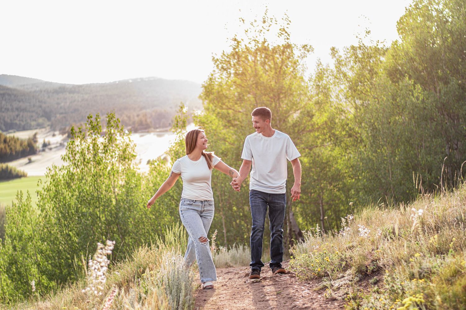 Couple holding hands walking on a scenic mountain trail surrounded by lush greenery and a lake in golden sunlight.