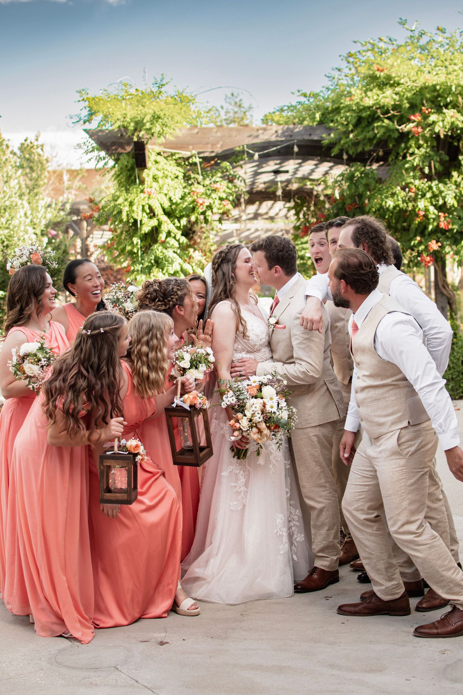 Bride and groom kiss surrounded by bridesmaids in coral dresses and groomsmen in tan suits at outdoor wedding.