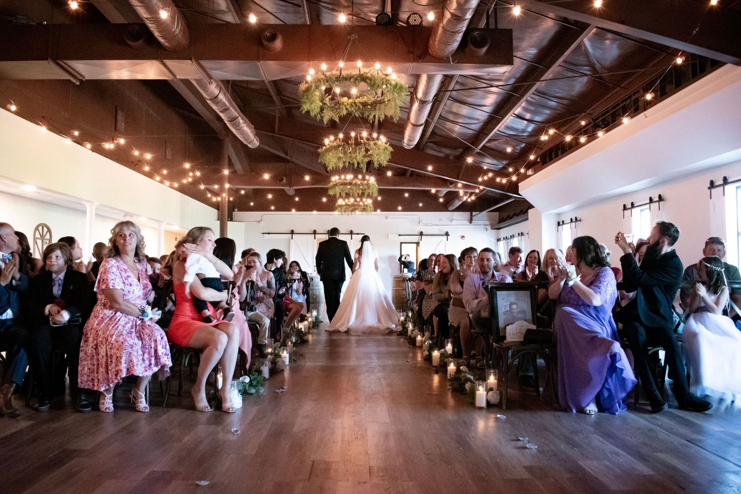 Guests watch a bride walk down the aisle at an elegant indoor wedding ceremony with string lights and wood beam ceiling.