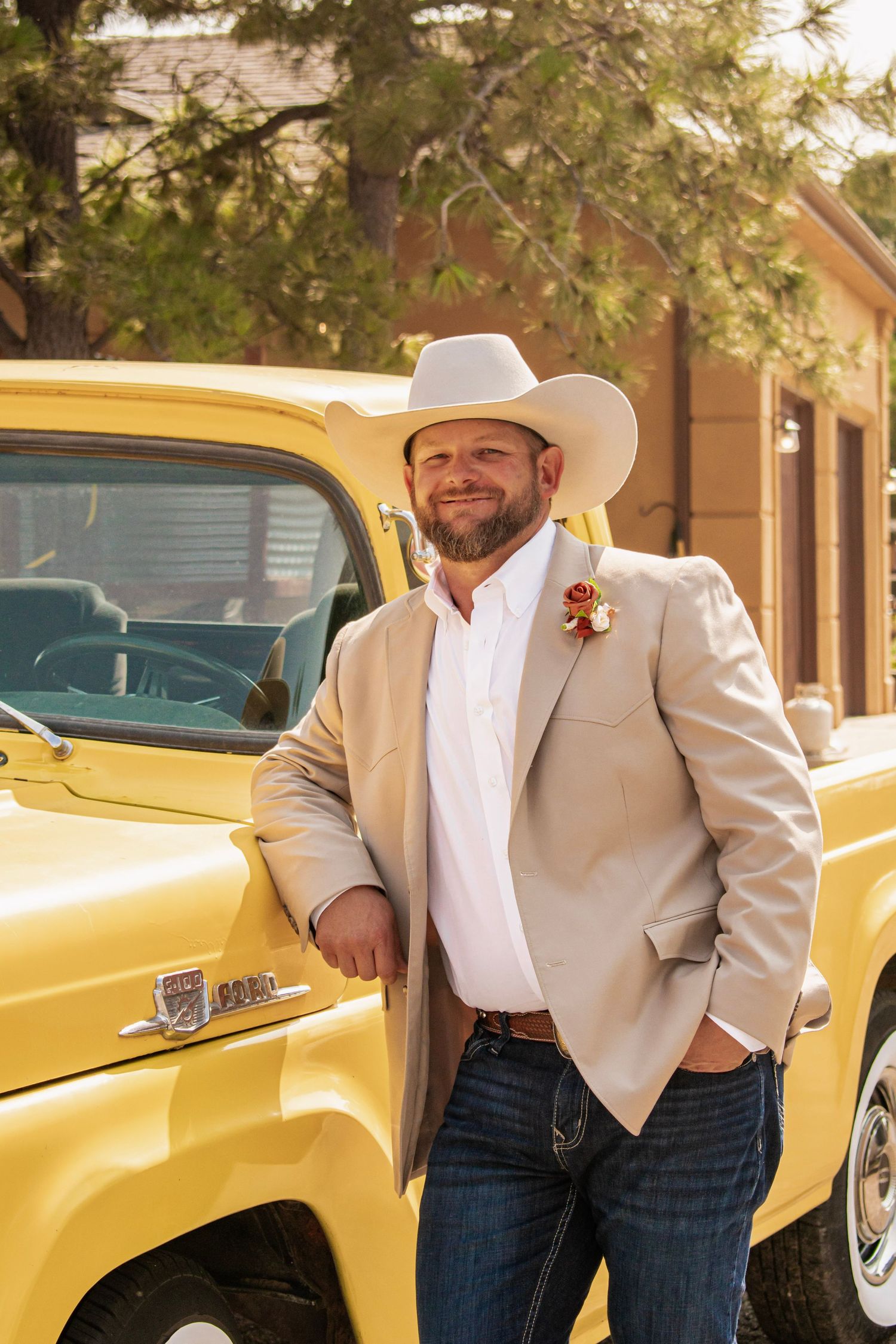 Man in white cowboy hat and tan blazer with boutonniere leaning against a classic yellow pickup truck.
