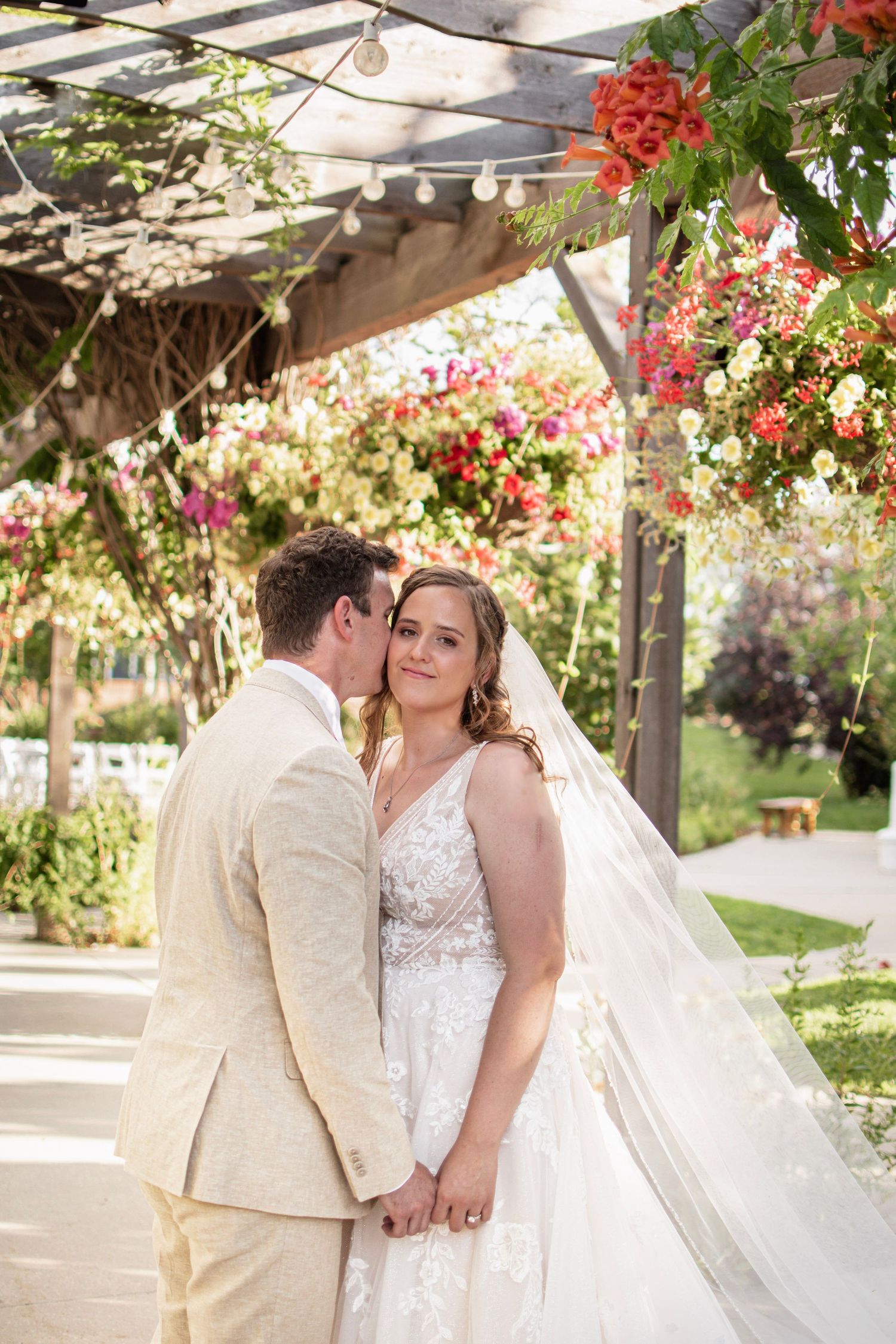 Bride and groom hold hands under a floral pergola, groom kissing bride's cheek at garden wedding, at Brookside Gardens,  Berthoud, CO
