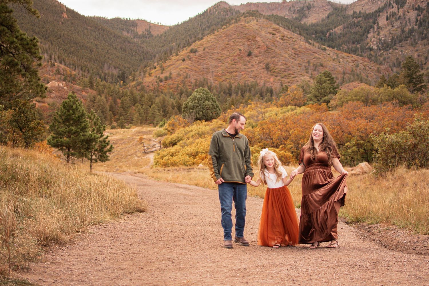 Family of three in coordinated fall outfits walking hand in hand along a dirt trail with autumn foliage and mountains in the background.