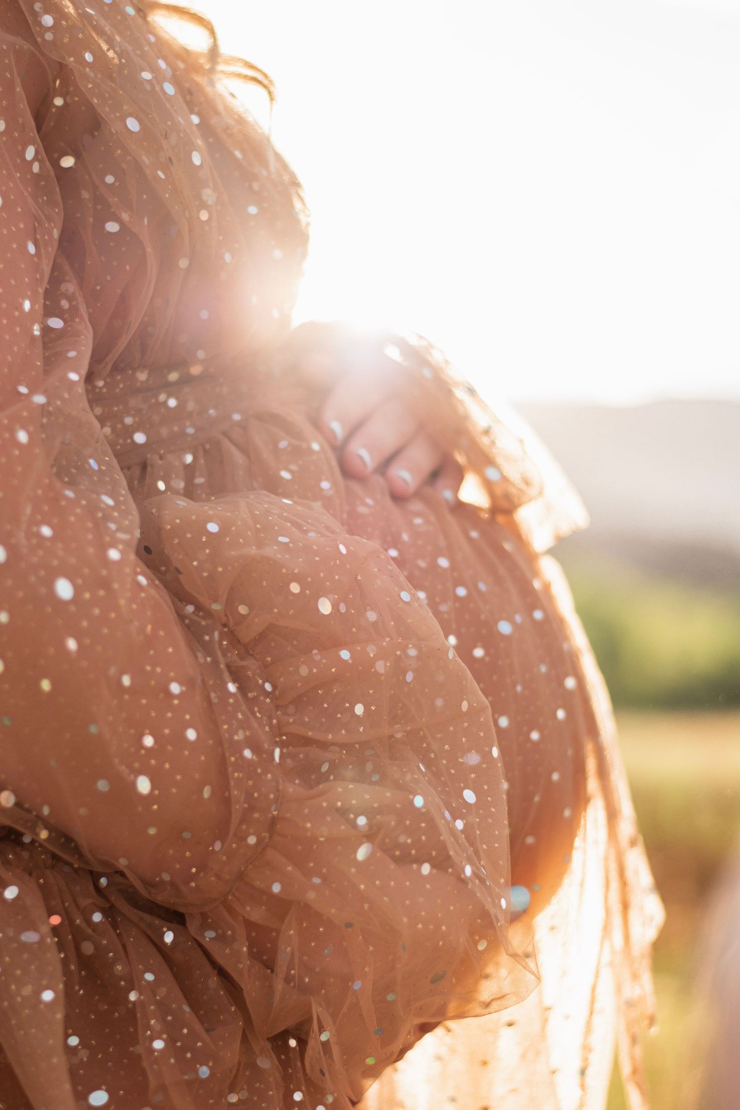 Pregnant woman in sheer polka dot dress cradling baby bump in golden sunlight outdoors.