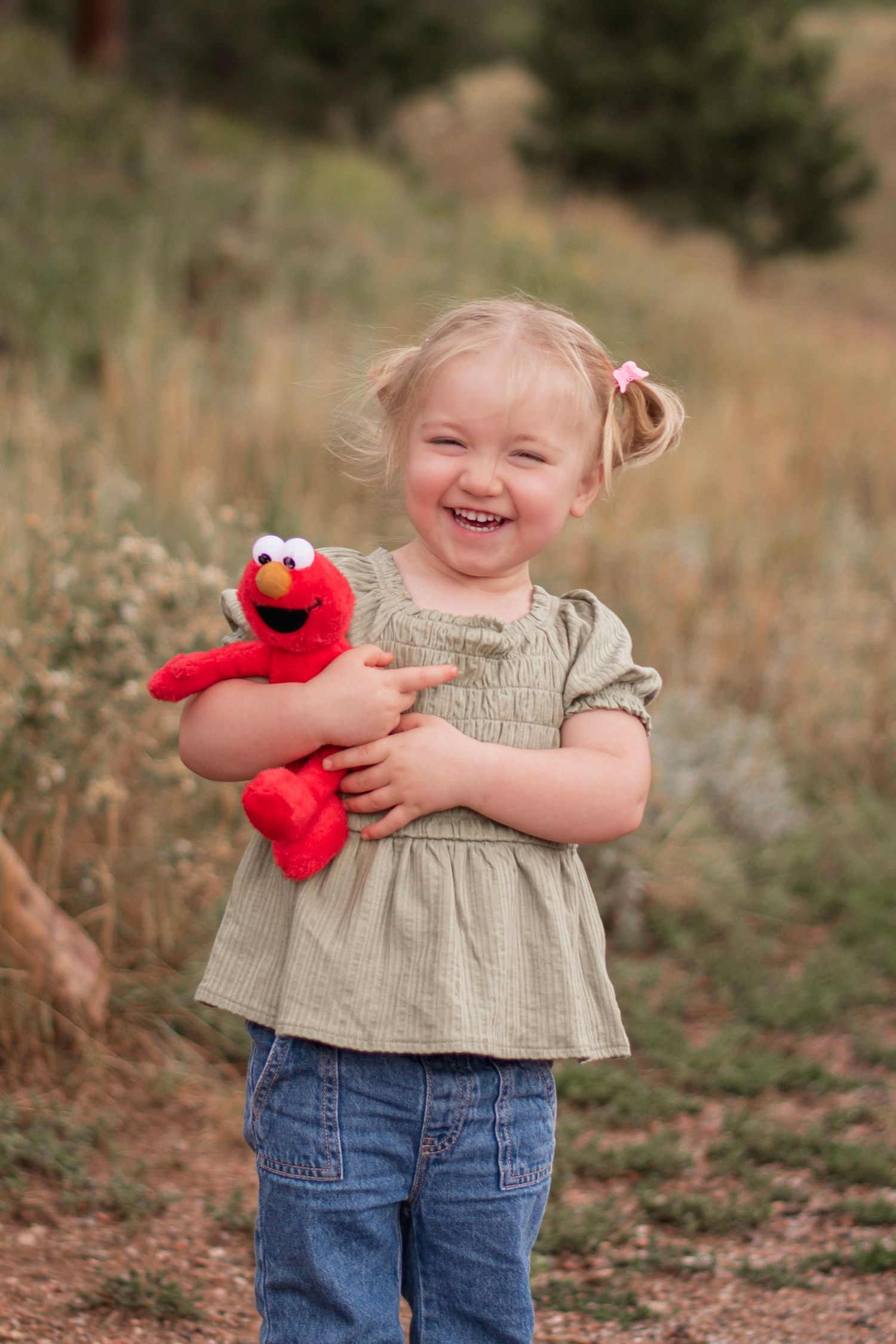 A laughing toddler girl with pigtails hugs an Elmo plush toy outdoors in a field, wearing a sage green top and jeans.