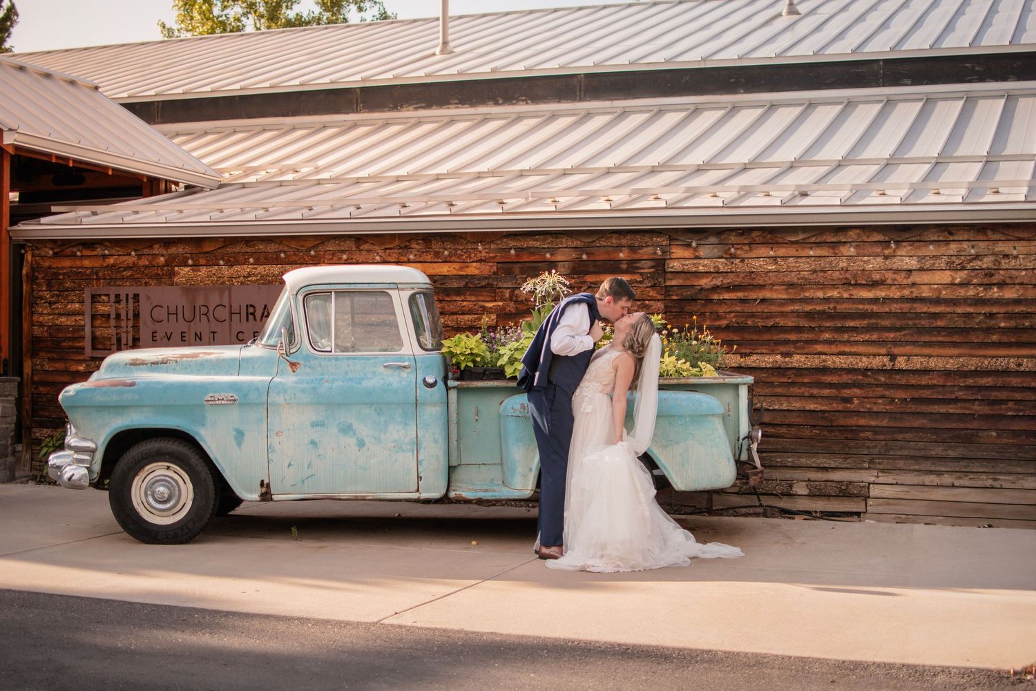 Bride and groom kissing beside a vintage turquoise pickup truck in front of a rustic wooden barn.
