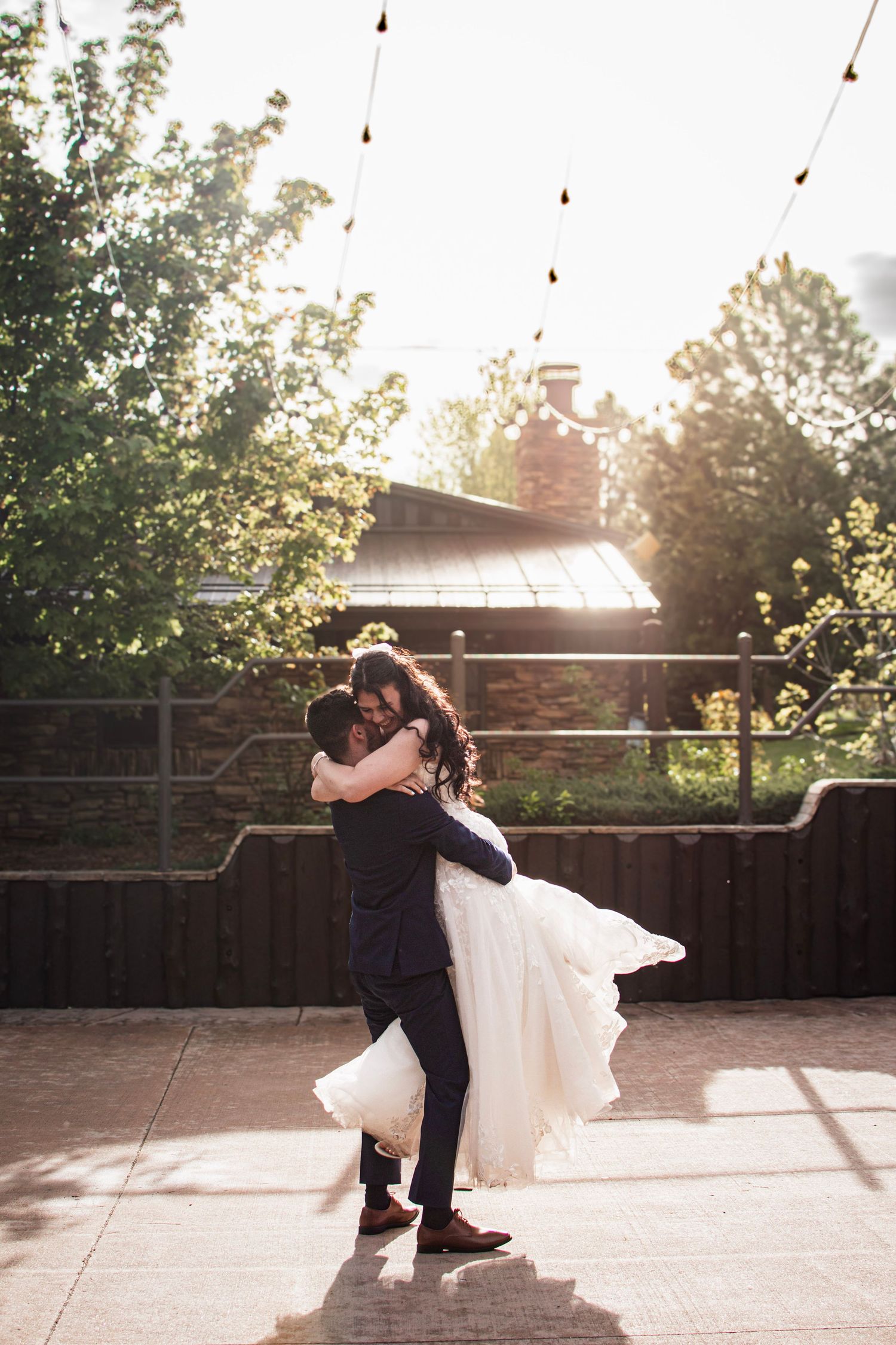 Groom lifting bride in white wedding dress outdoors on a sunny patio with string lights above.