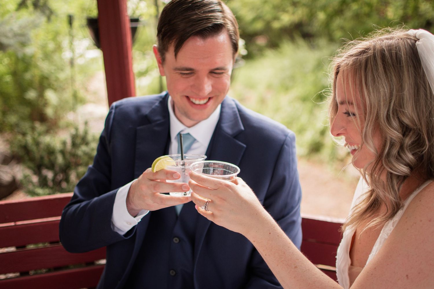 Smiling bride and groom in navy suit clink glasses on a red bench in a garden setting.