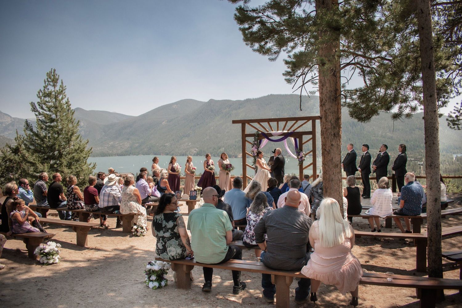 Outdoor mountain lakeside wedding ceremony with guests seated on wooden benches under pine trees at Grand Lake Lodge in Grand Lake, CO