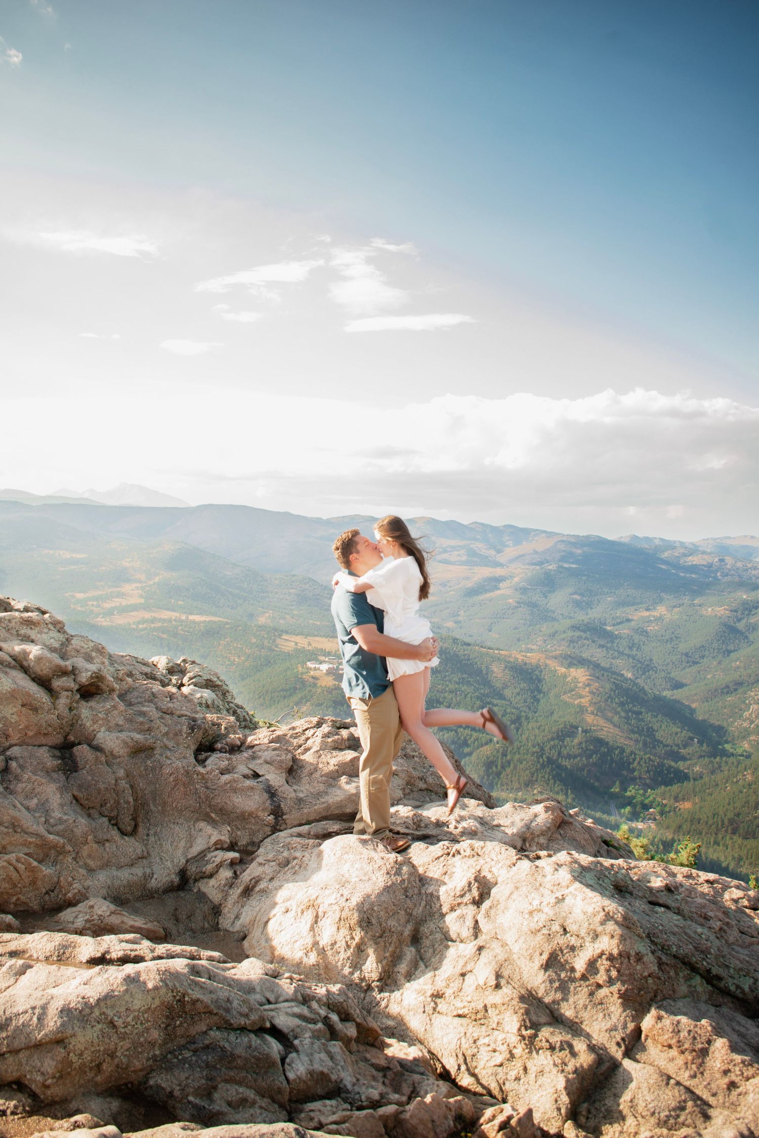 Couple embracing on rocky mountain summit with scenic green valley and blue-sky backdrop in Boulder, CO.