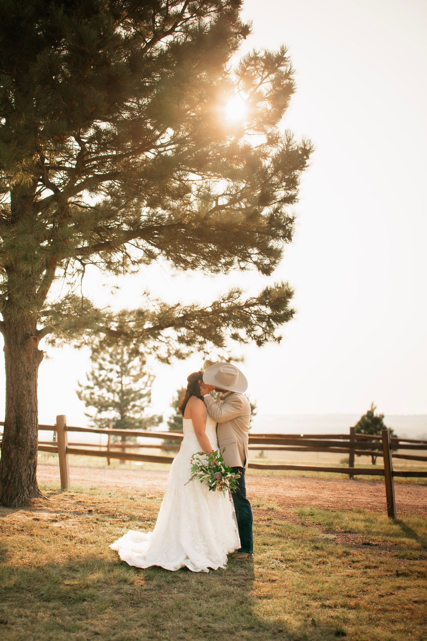 Bride and groom share a romantic kiss under a pine tree at golden hour on a rustic ranch wedding venue.