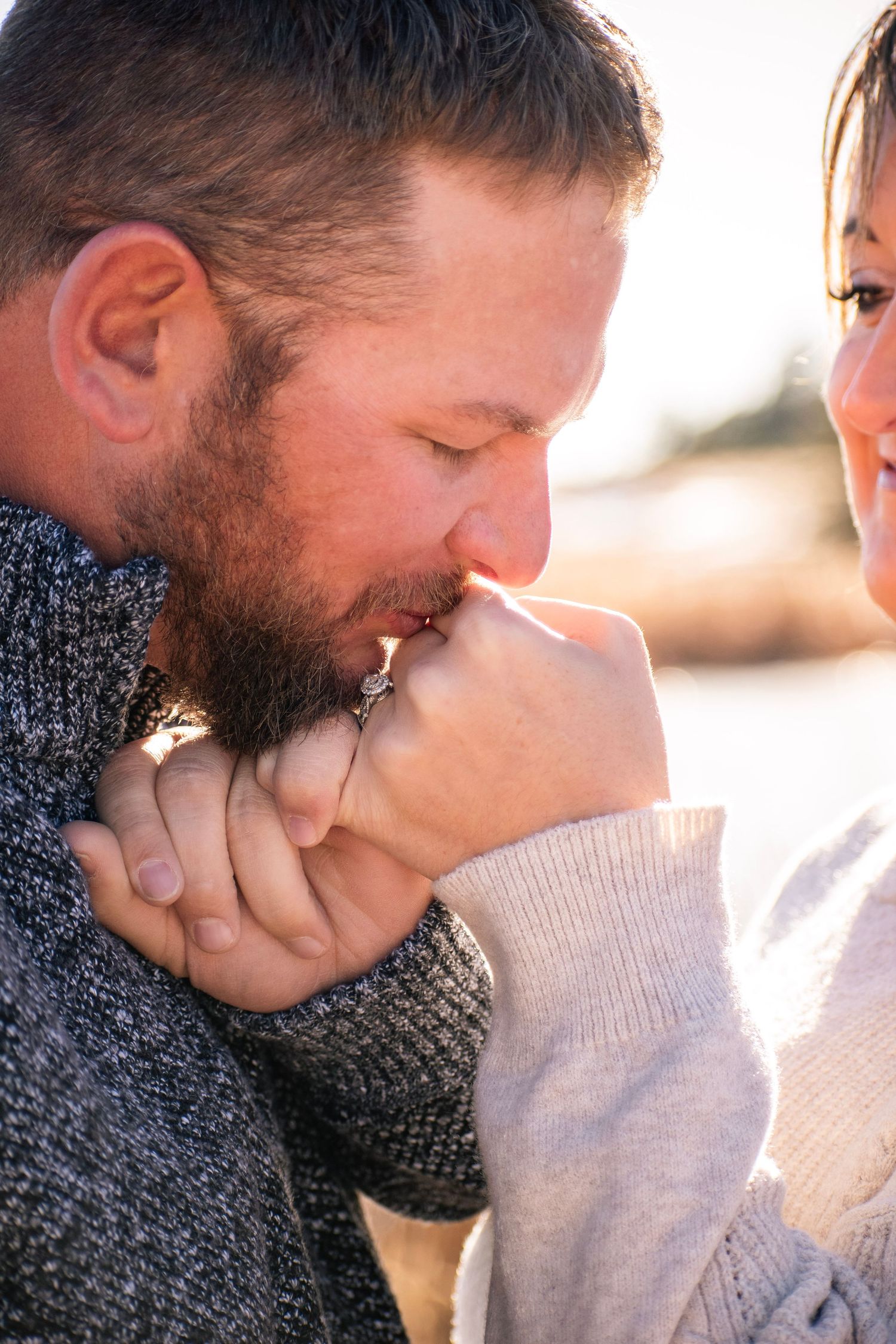 Man kissing woman's hand with engagement ring outdoors in warm sunlight, romantic couple moment.