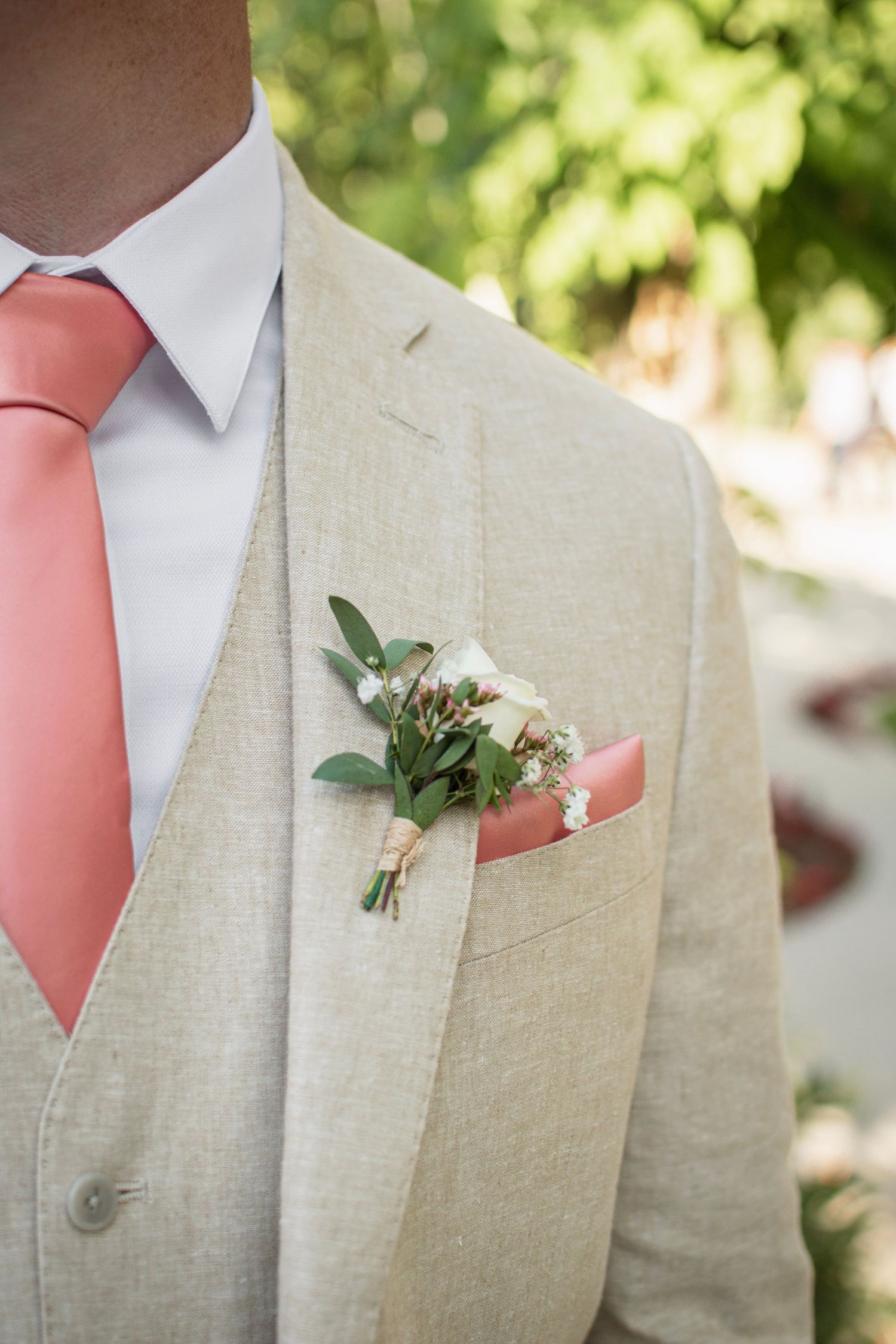 Groom in beige linen suit with coral tie, boutonniere of white flowers and greenery, and matching pocket square.