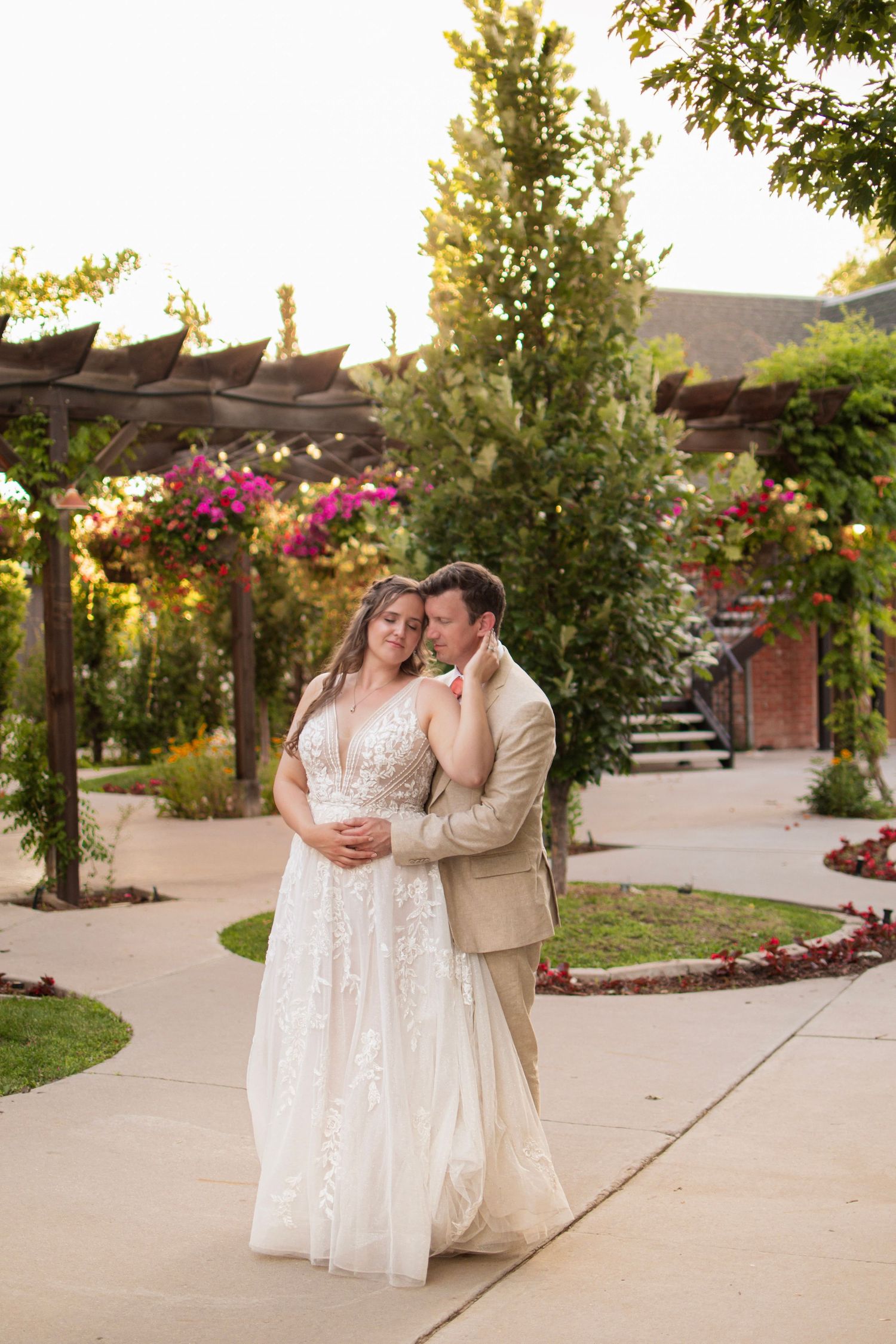 Bride and groom sharing a romantic moment on a garden pathway surrounded by colorful flowering pergolas at sunset.