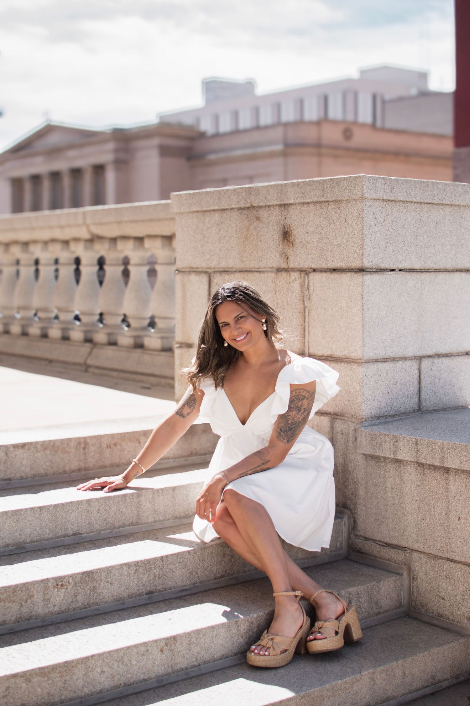 Smiling woman with tattoos in white dress sitting on stone steps outdoors near a classical building.