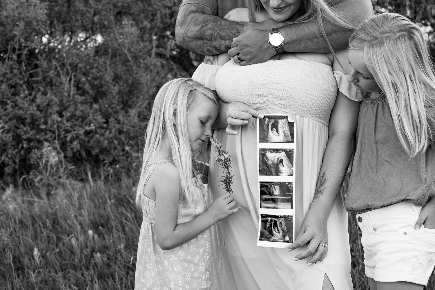 Black and white maternity photo of family holding ultrasound images outdoors, with two blonde girls embracing pregnant mother.