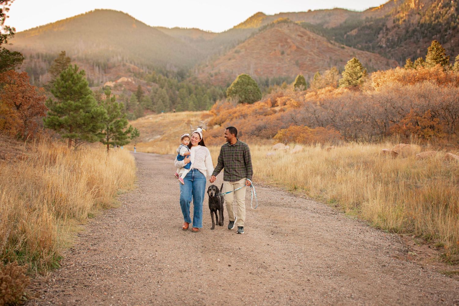 Family with baby and dog walking on a dirt trail through golden autumn fields with mountain backdrop at sunset at Blodgett Open Space.