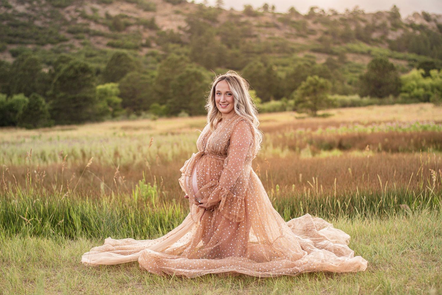 Pregnant woman in a flowing blush tulle maternity gown cradles her belly in a sunlit grassy field with rolling hills behind her.