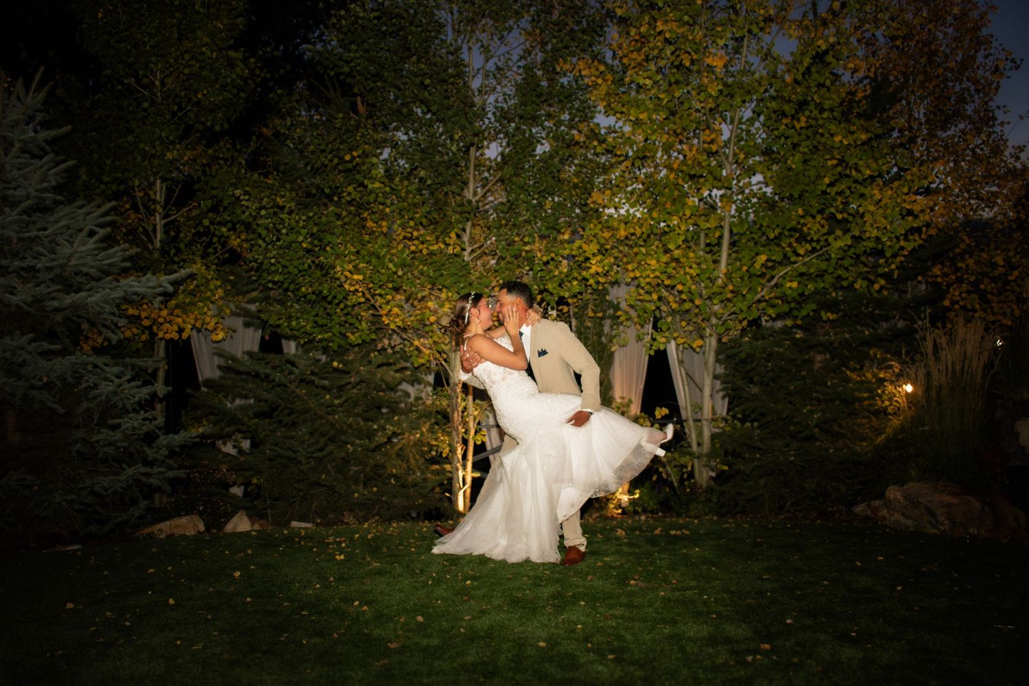 Groom lifts bride in a romantic nighttime wedding kiss surrounded by lush green trees in an outdoor garden setting.