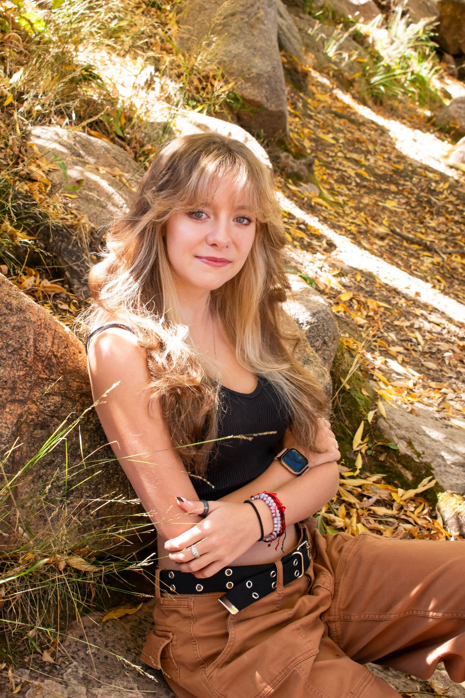 Young blonde woman sitting against rocks outdoors in autumn, wearing black top, brown pants, and colorful bracelets.