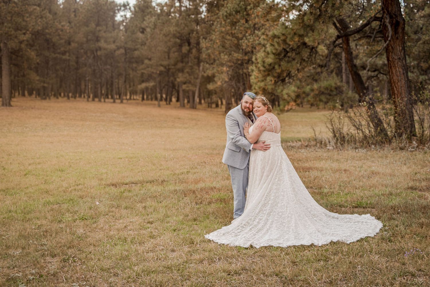 Bride and groom embracing in a rustic meadow surrounded by tall pine trees, bride wearing a lace gown with a long train.