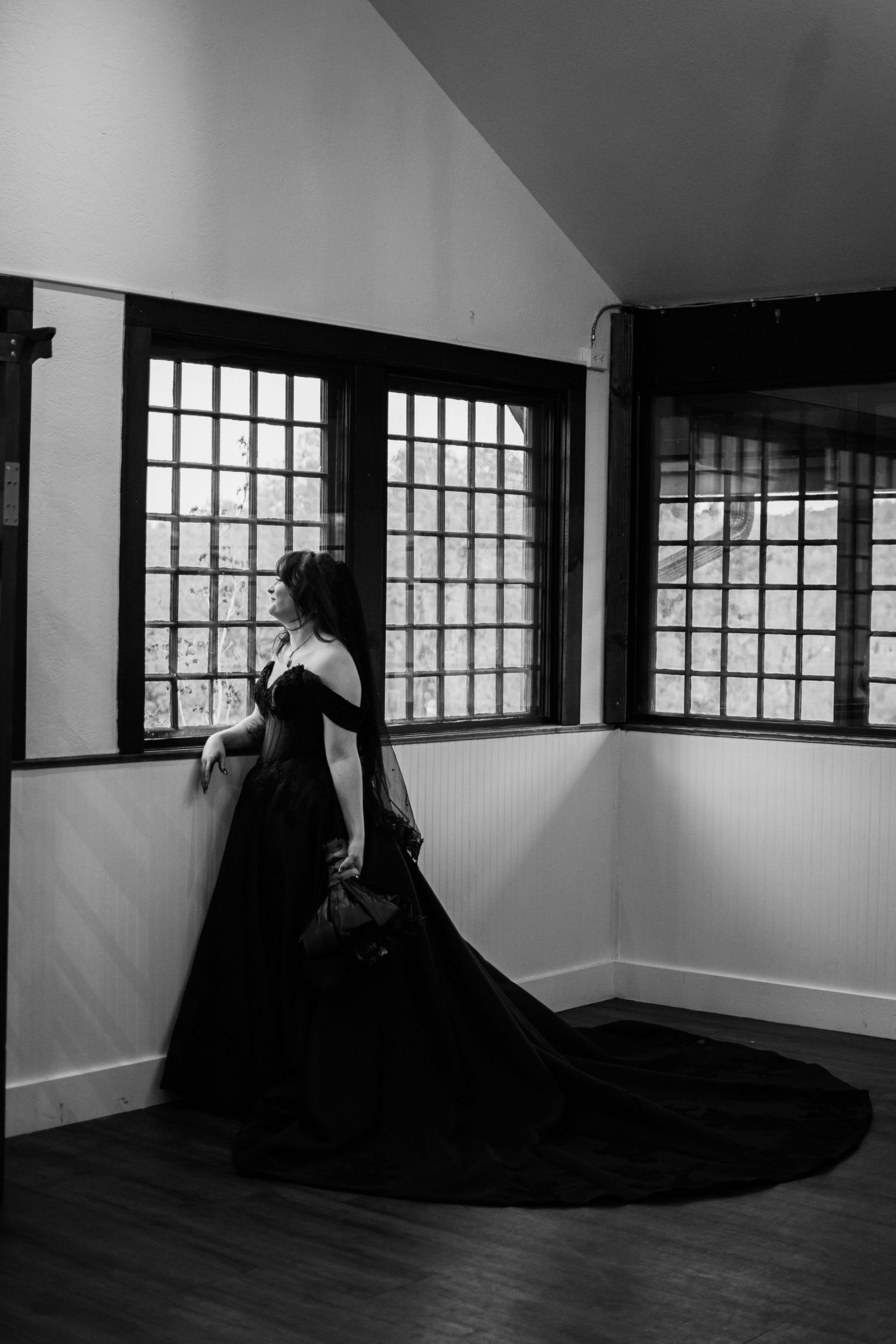Black and white photo of a woman in an elegant black ball gown with a long train standing by latticed windows.