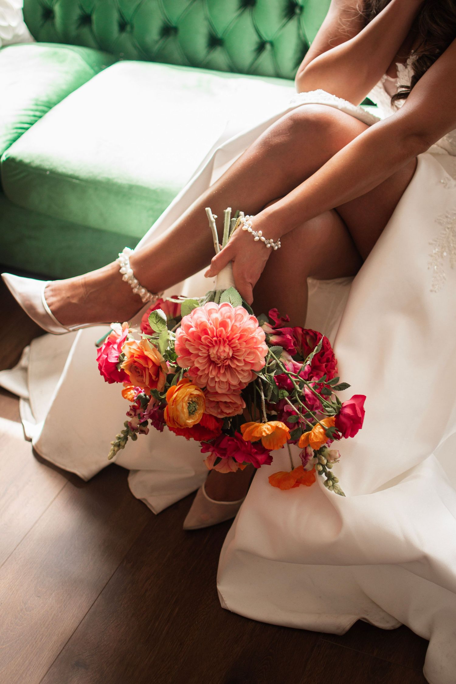 Bride in white dress and pearl heeled sandals sits on green velvet sofa holding vibrant pink and orange wedding bouquet.