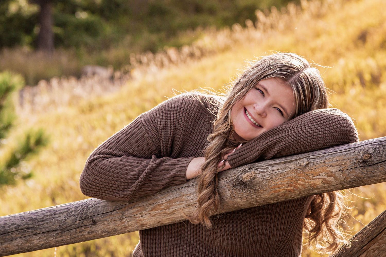 Smiling young woman in brown sweater resting on rustic wooden fence in golden autumn meadow.