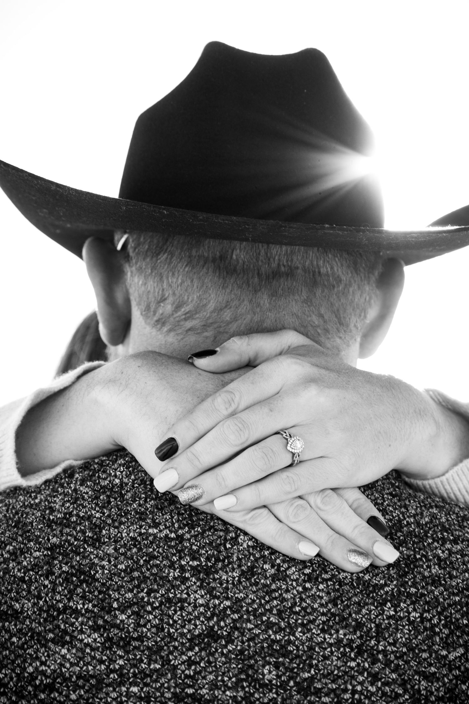 Black and white photo of couple embracing, woman's hand with diamond engagement ring resting on man's neck wearing cowboy hat.