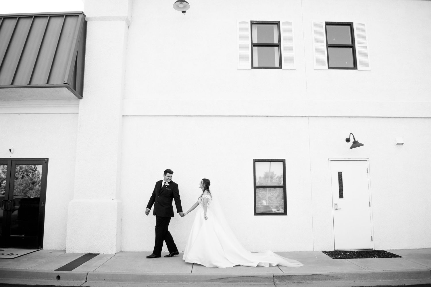 Black and white photo of bride and groom holding hands walking outside a white building at Creekside Terrace by Wedgewood