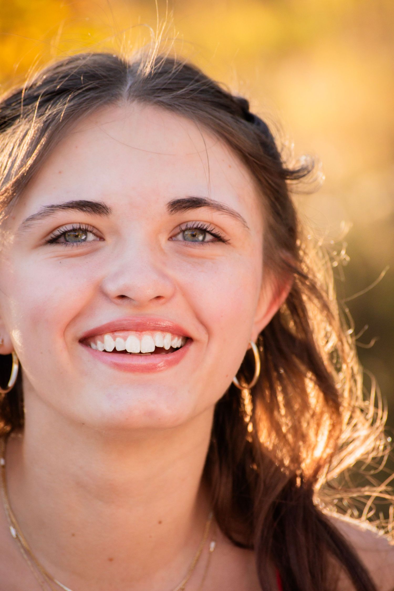 Smiling young woman with brown hair and hoop earrings outdoors in golden autumn sunlight.