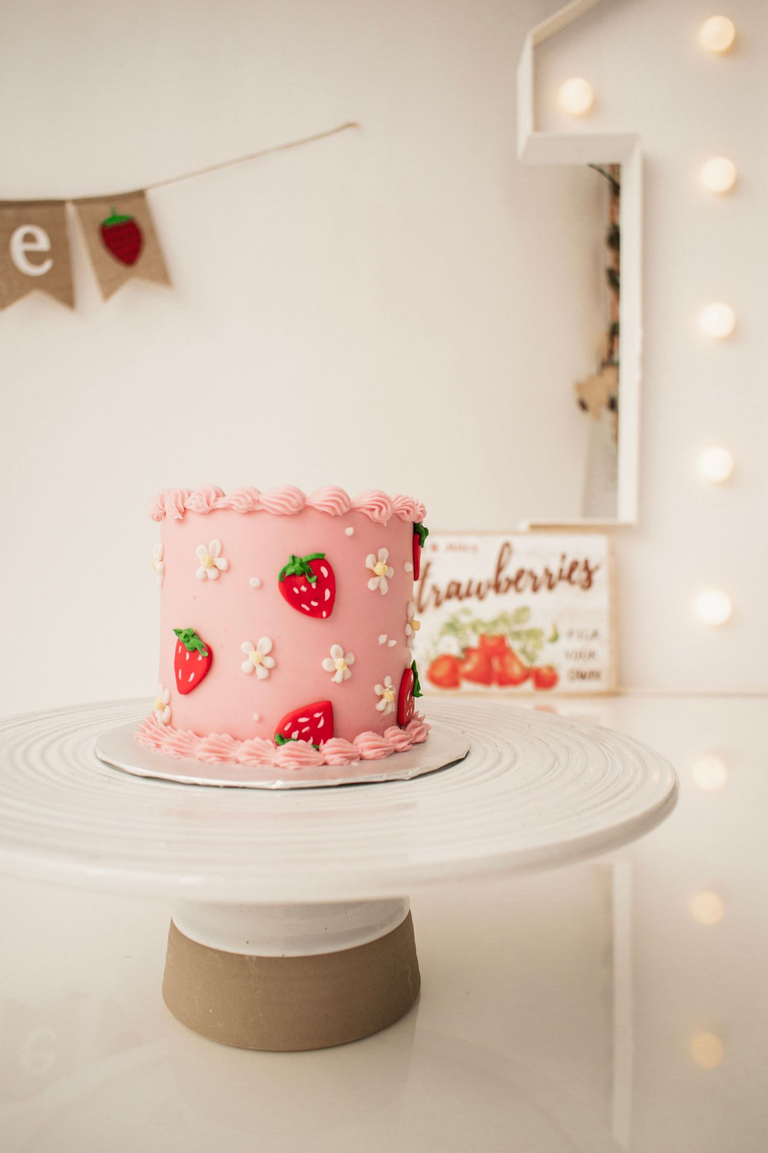 Pink strawberry birthday cake with red strawberry decorations on a white cake stand, first birthday party setup at Luminous Creative Studios